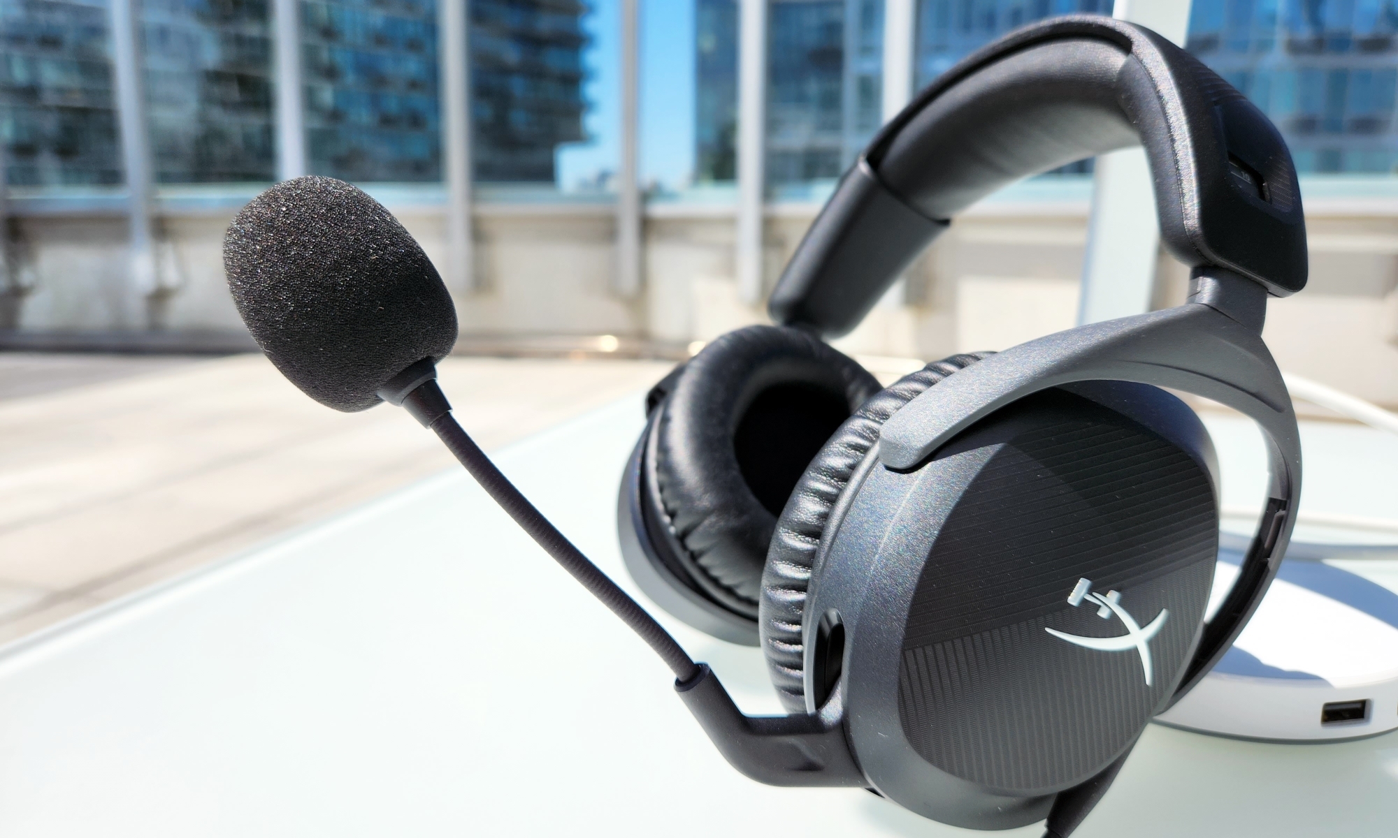 A black gaming headset with a built-in boom microphone, the HyperX Cloud Stinger 2, rests on a white table in an outdoors setting. | DeviceDaily.com